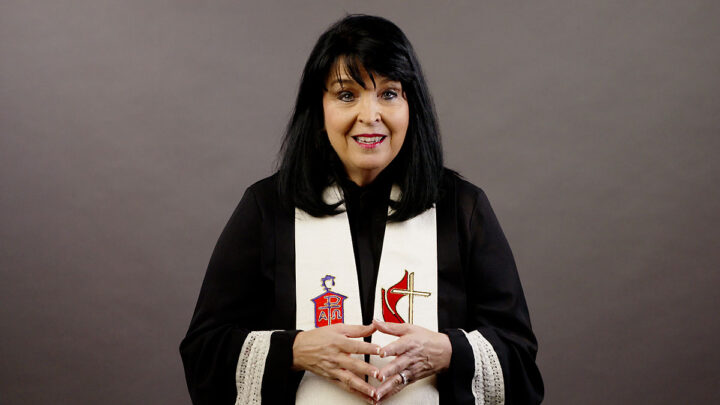 Bishop Connie Mitchell Shelton dressed in a robe with a white stole sitting in front of a gray background