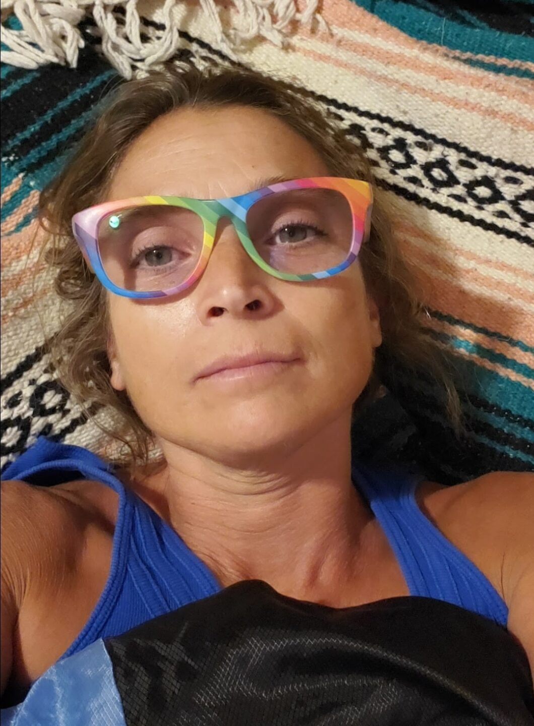 middle-aged female, no jewelry, emotionally neutral expression, cafe au lait skin color, green eyes, brown curly shoulder length hair, wearing large lens eyeglasses with rainbow color frames and a royal blue tank top
