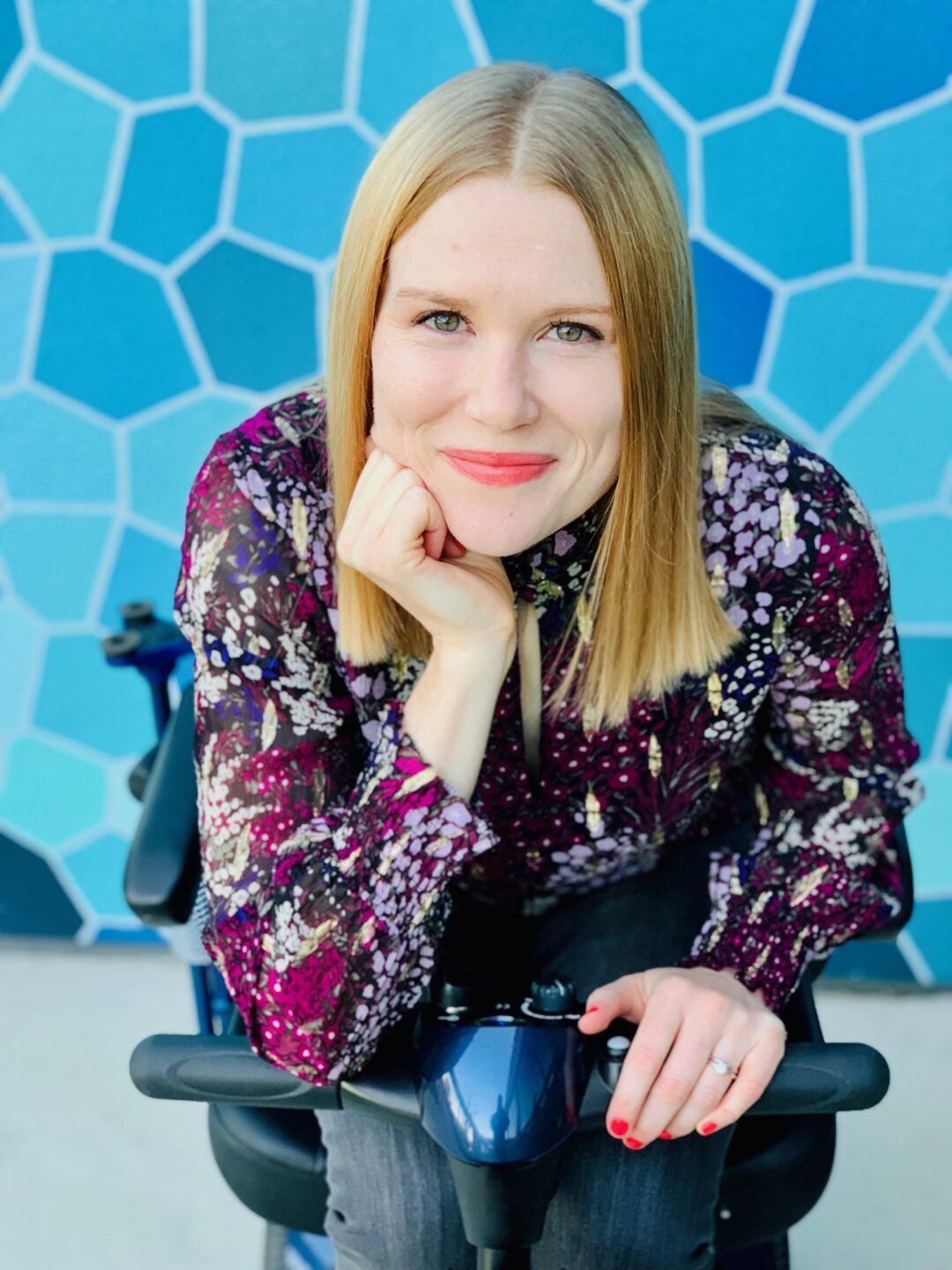 a white woman with blonde hair wearing a purple blouse and sitting in her mobility scooter in front of a wall with blue geometric shapes
